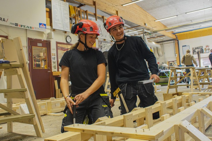 Två personer med röda hjälmar arbetar med träkonstruktioner i en verkstadsmiljö.