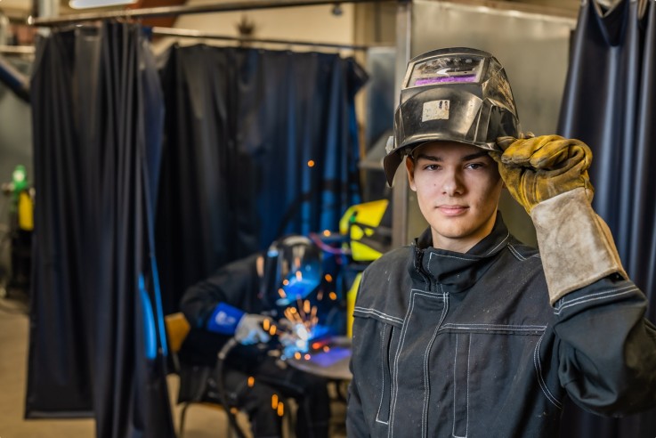 Två personer i svetsverkstad, båda bär skyddsutrustning och svetsmask, en arbetar och en tittar mot kameran.