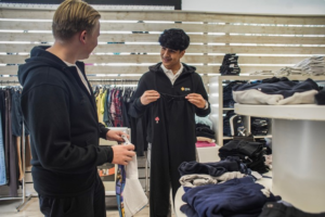 Två personer tittar på kläder i en butik, en håller upp ett svart plagg, omgivna av hyllor och klädhögar.