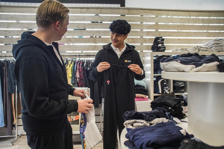 Två personer tittar på kläder i en butik, en håller upp ett svart plagg, omgivna av hyllor och klädhögar.