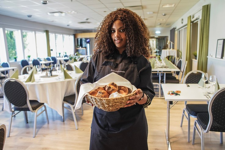 En person i svart förkläde håller en korg med bröd i en ljus, dukad matsal.