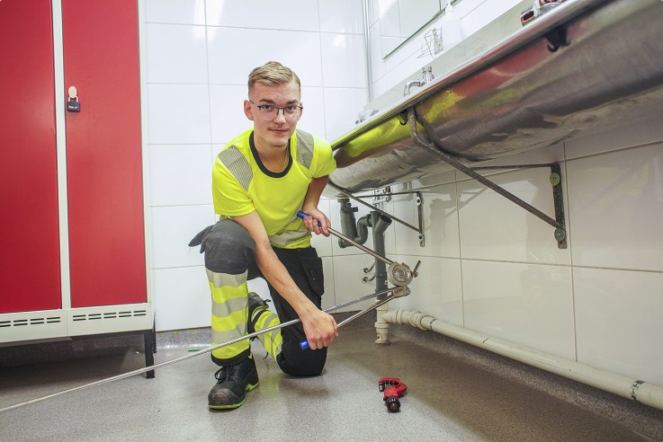 En person i arbetskläder arbetar med ett verktyg under ett handfat i ett kaklat badrum.