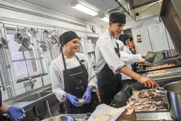 Flera personer i kockkläder lagar mat tillsammans i ett storkök med rostfria ytor och köksredskap.