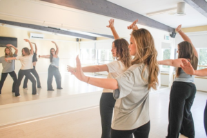 Fyra personer övar dans i en ljus dansstudio med spegelvägg, alla har armarna uppsträckta.