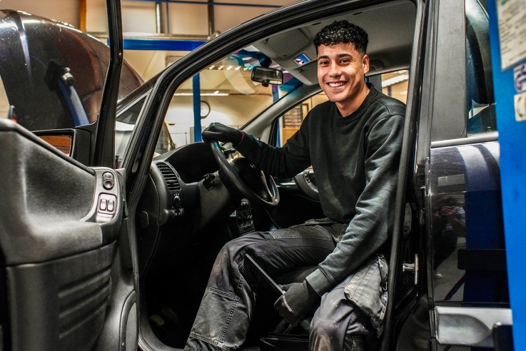 En mekaniker sitter i förarsätet på en bil i en verkstad, med dörren öppen och verktyg i handen.