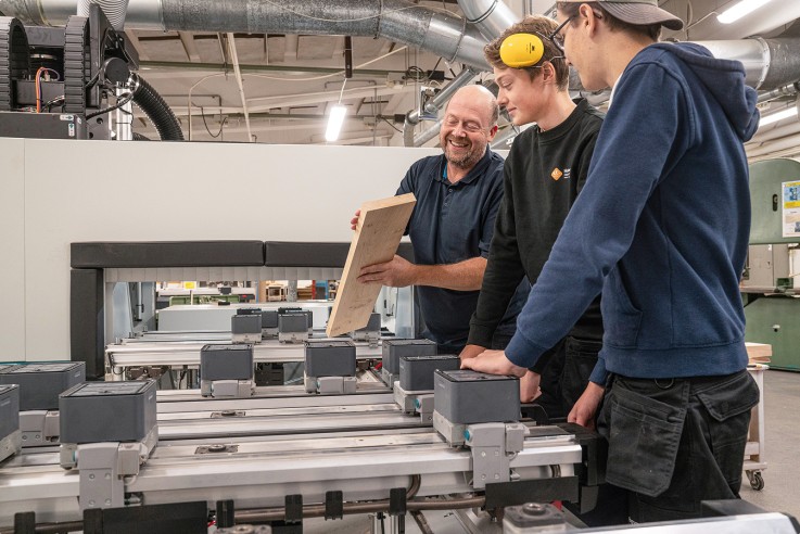 Tre personer arbetar vid en industriell maskin, en håller en träplanka och instruerar de andra.