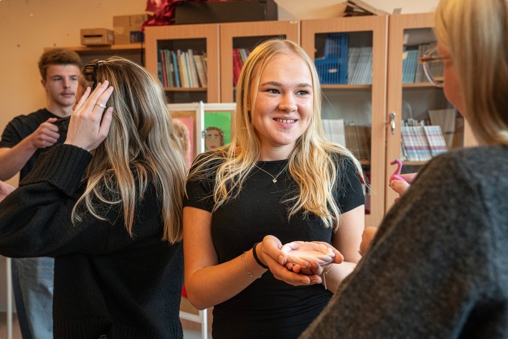 Fyra personer står i ett klassrum, två kvinnor i fokus, en håller något i händerna. Bokhyllor syns i bakgrunden.