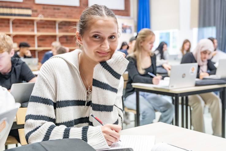 Studenter sitter i ett klassrum och arbetar vid sina bänkar med datorer och anteckningsblock.
