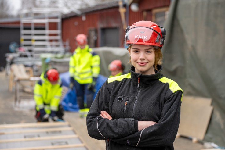Person i arbetskläder och hjälm står med armarna i kors på en byggarbetsplats, fler personer syns i bakgrunden.