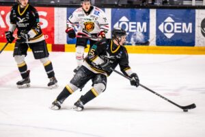 Ishockeyspelare i svart och vitt spelar match på isen, pucken syns vid en klubba.
