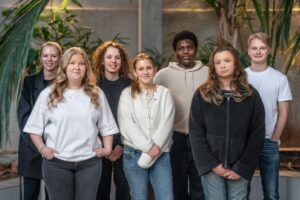 En grupp på sju personer står tillsammans inomhus bland gröna växter. Ansiktena är suddade.