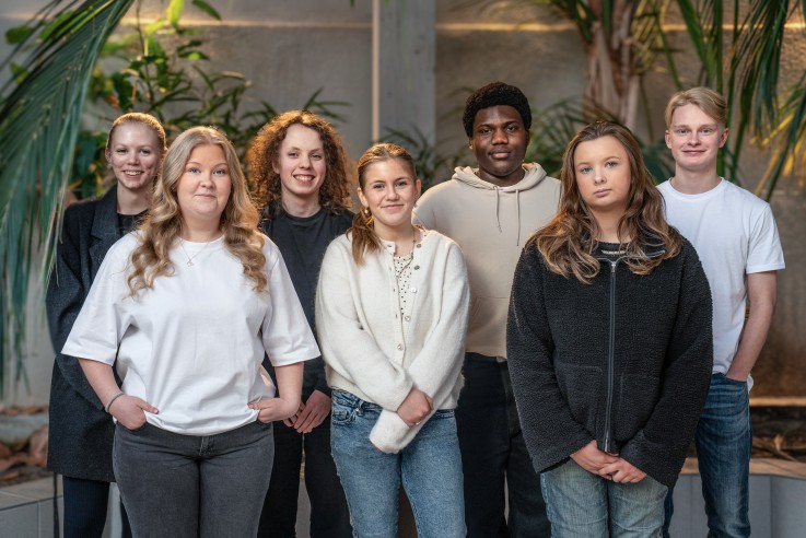 En grupp på sju personer står tillsammans inomhus bland gröna växter. Ansiktena är suddade.