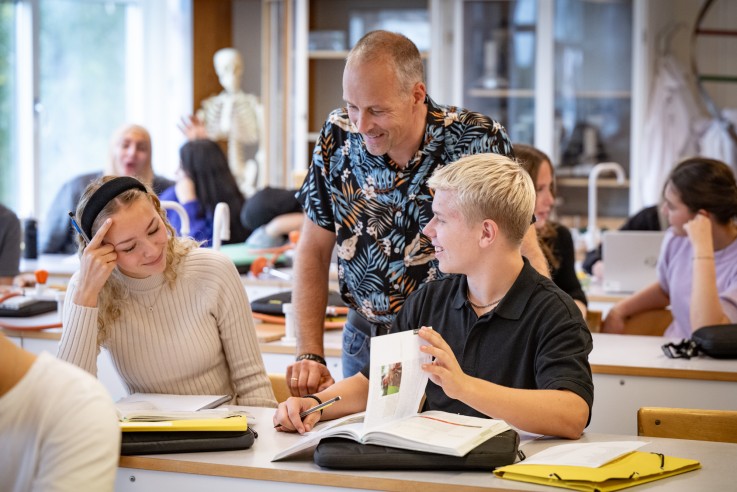 Lärare hjälper två elever i ett klassrum med böcker och anteckningar på bänken.