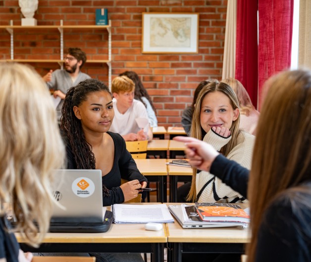 En grupp elever sitter vid bänkar i ett klassrum och diskuterar, med datorer och böcker framför sig.