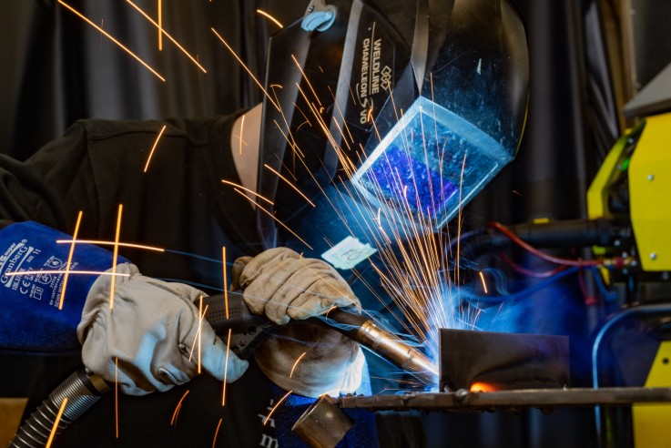 En person svetsar metall med skyddshjälm och handskar, gnistor flyger från arbetsytan.