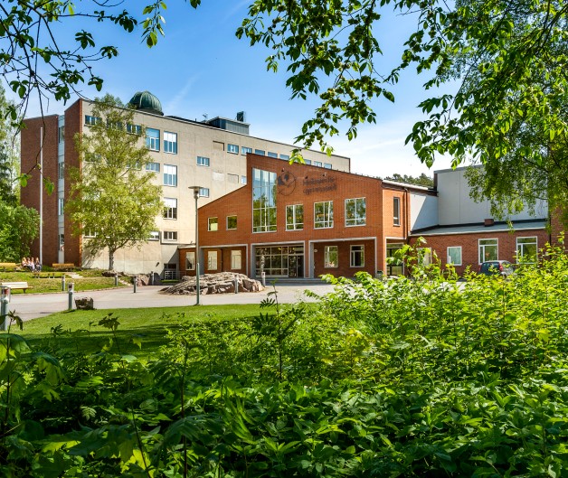 En skolbyggnad i tegel och betong omgiven av grönska och träd en solig dag.