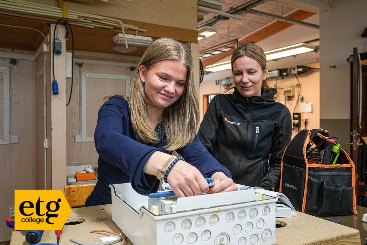 Två personer arbetar med elinstallation i ett klassrum, verktyg och elskåp syns på bordet.