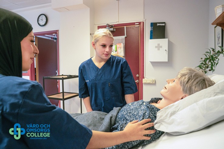 Tre personer i vårdkläder står vid en säng i ett vårdrum, en person ligger ner och får omvårdnad.