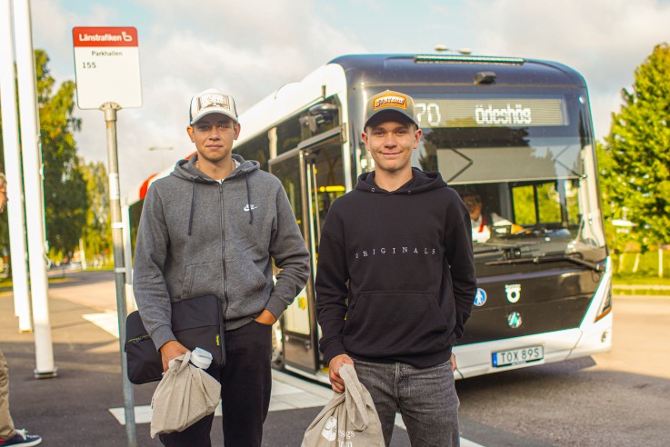 Två personer står vid en busshållplats framför en buss, båda håller tygpåsar och bär kepsar.