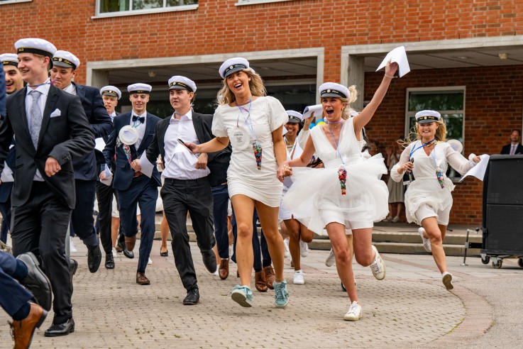 Studenter i vita kläder och mössor springer glada ut från en skolbyggnad vid examen.