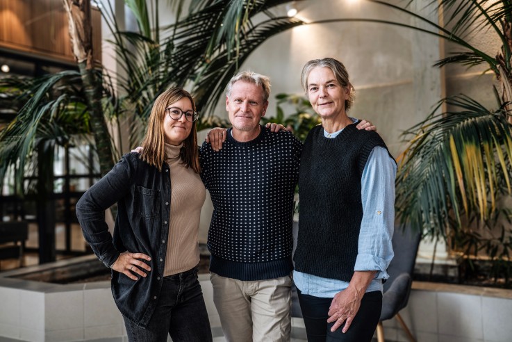 Tre personer står tillsammans inomhus bland stora gröna växter, med armarna om varandra.