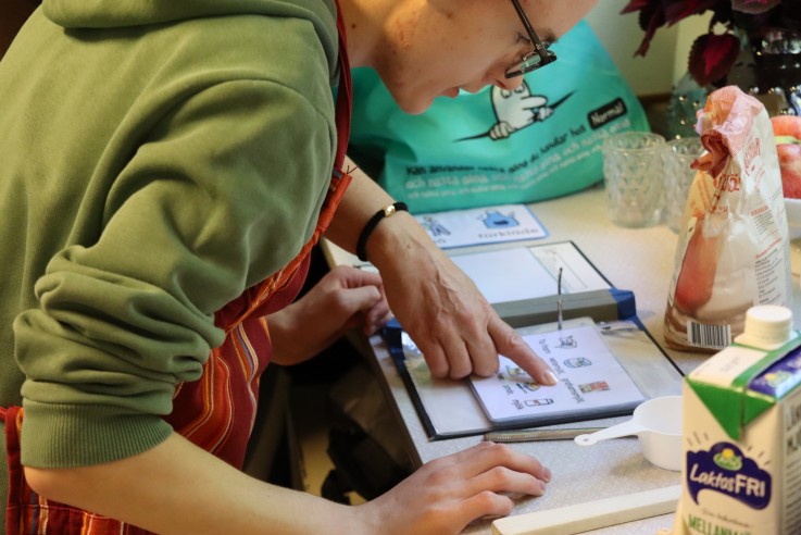 Person i grön tröja och randigt förkläde pekar på ett receptblad i köket, omgiven av matvaror.