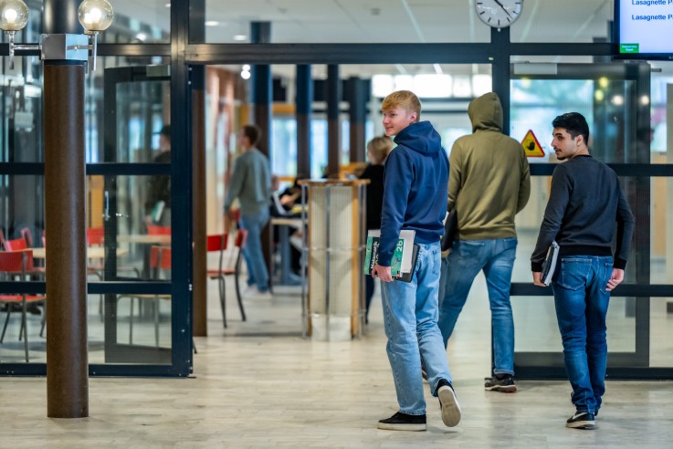 Tre unga personer går i en skolmiljö med böcker i händerna, suddiga ansikten för anonymitet.