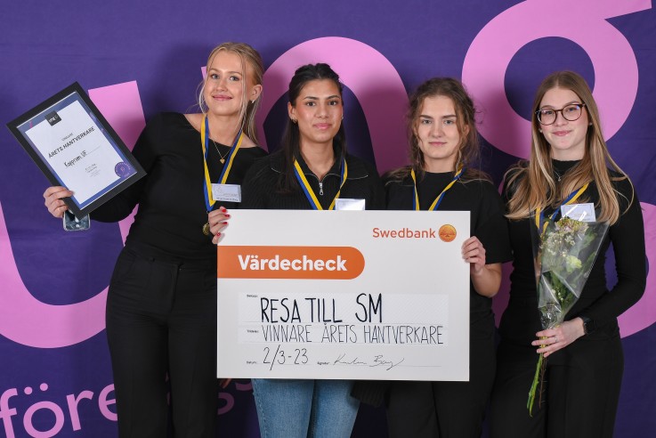 Fyra personer poserar med diplom, blommor och en stor symbolisk check framför en lila bakgrund.