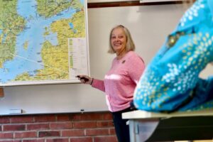 Lärare pekar på en karta i klassrum medan en elev sitter vid en bänk i förgrunden.