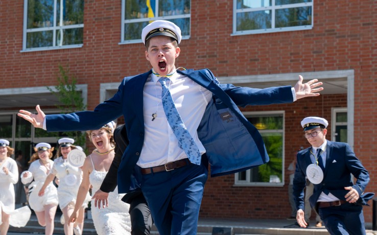 Studenter i vita mössor och festkläder springer glada ut från en skolbyggnad.