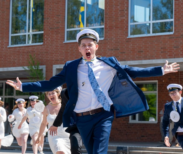 Studenter i vita mössor och festkläder springer glada ut från en skolbyggnad.