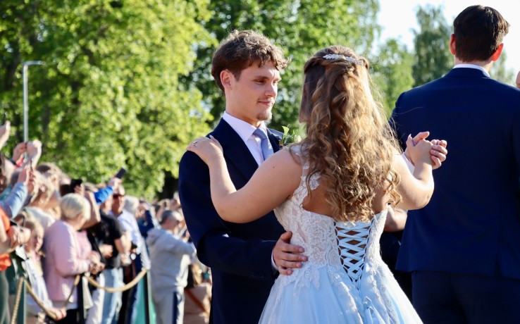 Ett par dansar utomhus i festkläder inför en publik, med grönskande träd i bakgrunden.