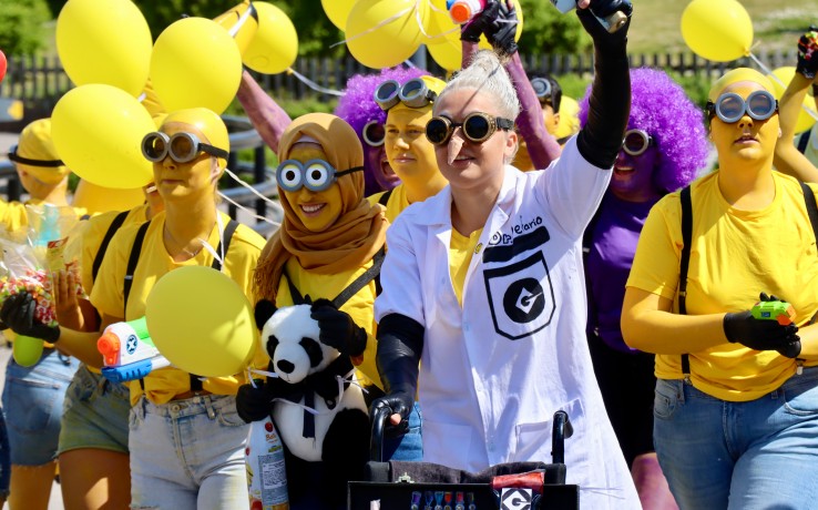 Grupp människor utklädda i gula kläder och labbrock, håller gula ballonger och leksaker under en utomhusparad.