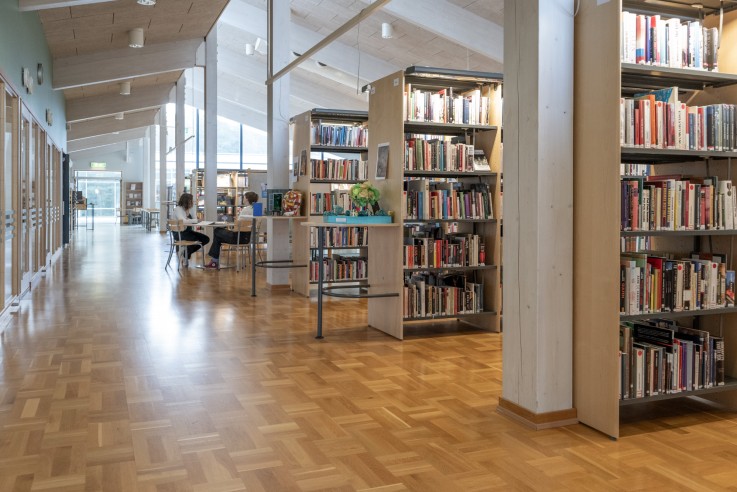 Ljust bibliotek med trähyllor, böcker och två personer som sitter och studerar vid ett bord.