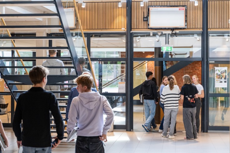 Ett gäng ungdomar står och går i en ljus skolmiljö med trappa och glasväggar.