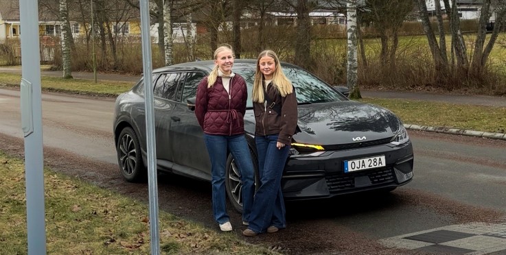 Två personer står framför en grå bil vid ett övergångsställe på en landsväg, med ett övergångsställeskylt synlig.