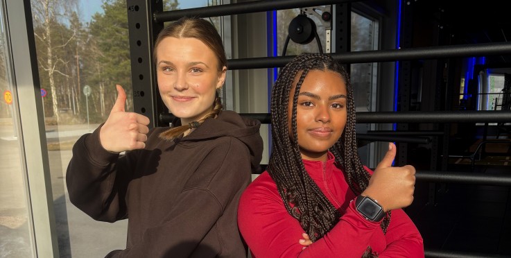 Två personer står i ett gym, ger tummen upp och poserar framför ett klätterställning.