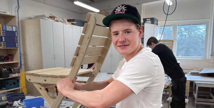 Två personer arbetar med träslöjd i ett ljust klassrum, en håller i en trästol vid arbetsbordet.