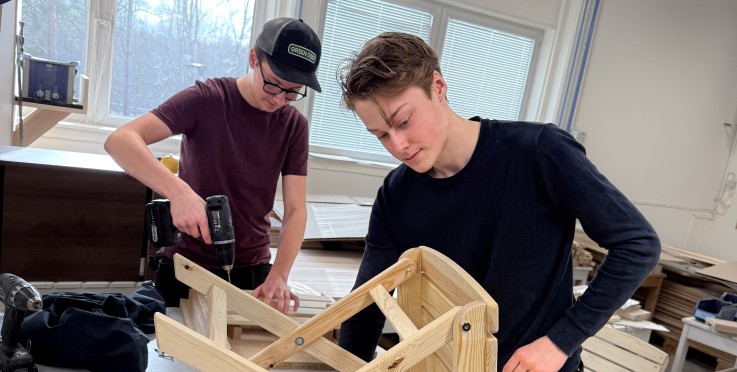 Två personer bygger en trästol i ett ljust slöjdrum med verktyg på bordet.