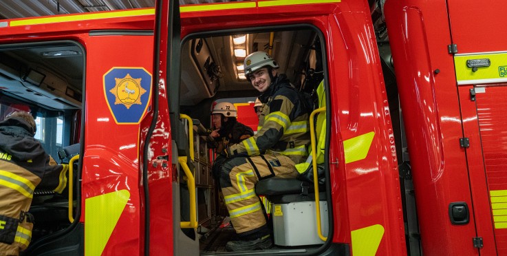 Brandmän sitter i en röd brandbil med dörren öppen, redo för utryckning inomhus.