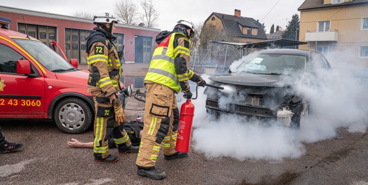Brandmän släcker en bilbrand med brandsläckare på en gata, rök väller ut från motorhuven.