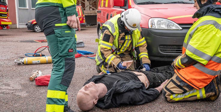Räddningspersonal övar första hjälpen på en docka utomhus vid en röd utryckningsbil.