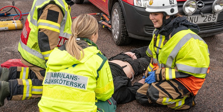 Räddningspersonal och ambulanssjuksköterska hjälper en person på marken vid en bilolycka.