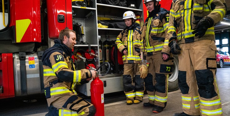 Fyra brandmän i skyddskläder står vid en brandbil och hanterar en brandsläckare inomhus.