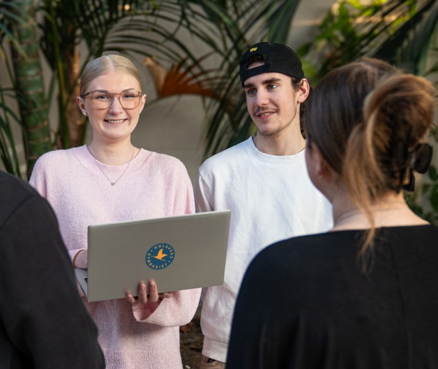 Fyra personer står i en grupp inomhus bland stora gröna växter, en håller en laptop.