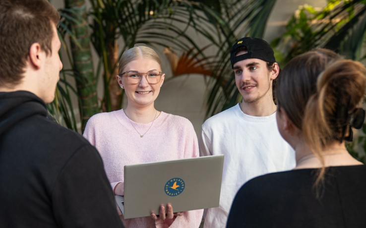 Fyra personer står i en grupp inomhus bland stora gröna växter, en håller en laptop.