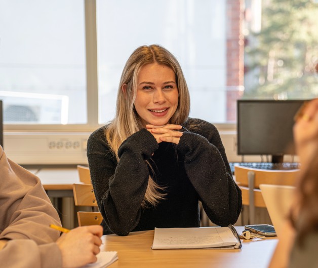 Tre personer sitter runt ett bord i ett klassrum och diskuterar, anteckningsblock och datorer syns.
