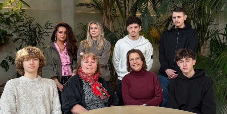 En grupp på åtta personer sitter och står runt ett runt bord i ett rum med stora gröna växter.