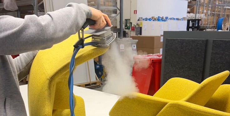 En person ångar en gul tygstol med en handhållen steamer i en industriell miljö.