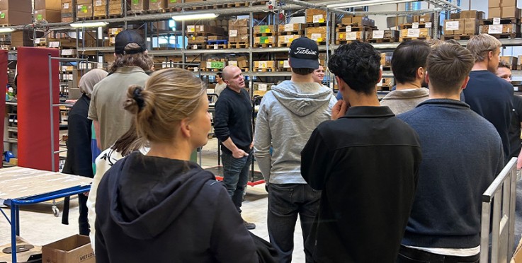 En grupp människor står i ett lager och lyssnar på en person som talar.